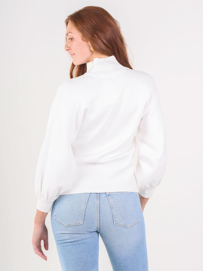 A woman with long red hair wears the Lolli Sweater, a sustainable white tunic with long sleeves and a high neck, paired with light blue jeans. She stands against a plain white background, looking over her left shoulder.