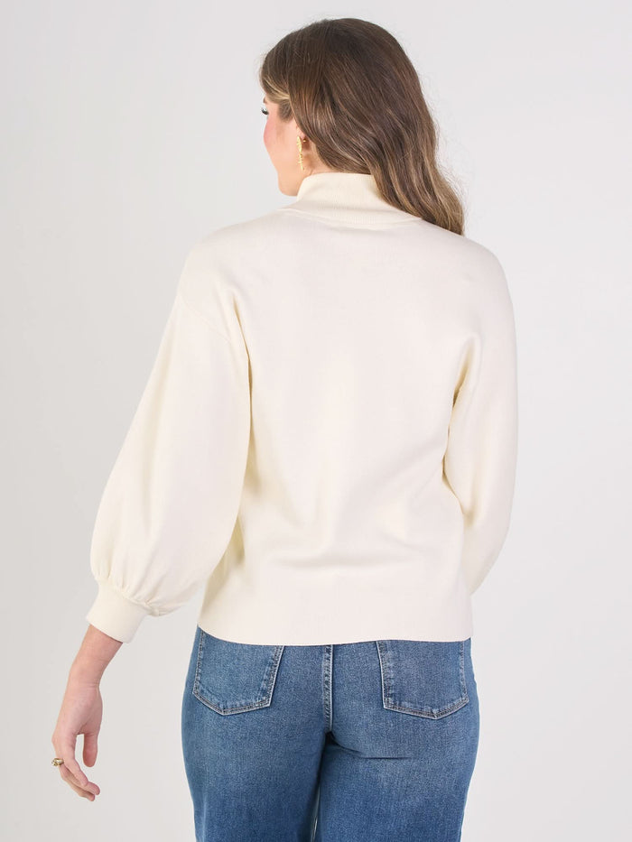A woman stands with her back facing the camera, wearing the Lolli Sweater in cream with a stand collar and blouson sleeves with fitted cuffs, paired with blue jeans, her long wavy hair cascading against a light background.