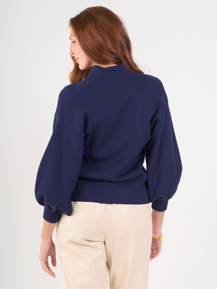 A woman with long red hair stands facing away, wearing a sustainable Lolli Sweater with balloon sleeves in navy blue and light beige pants against a plain white background.