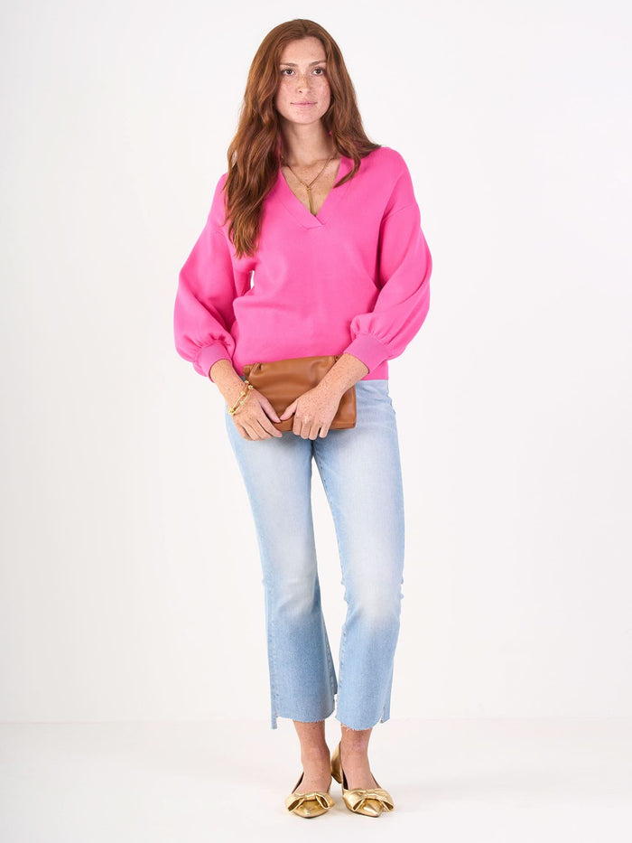 A woman with long red hair wears the Lolli Sweater with blouson sleeves, paired with light blue cropped jeans and gold shoes. She holds a tan clutch bag and stands against a plain white background.