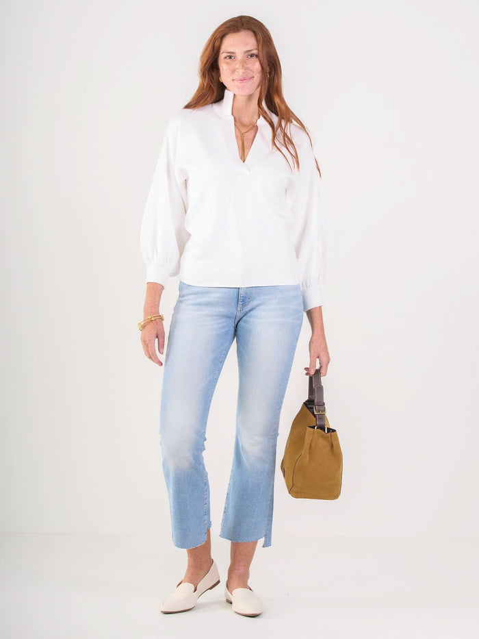 A woman with long red hair stands against a plain background, smiling gently. She wears the Lolli Sweater with light blue cropped jeans, white shoes, and holds a tan handbag in her left hand.