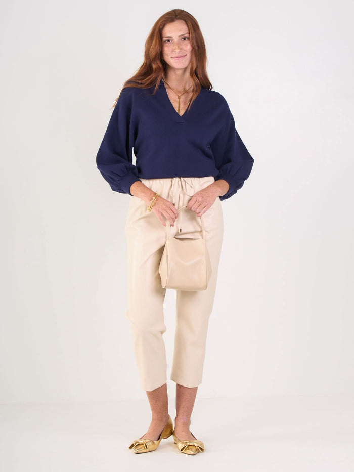 A woman with long auburn hair wears the navy Lolli Sweater, beige cropped pants, and gold flats. She holds a small beige handbag and stands against a plain white background, smiling softly.