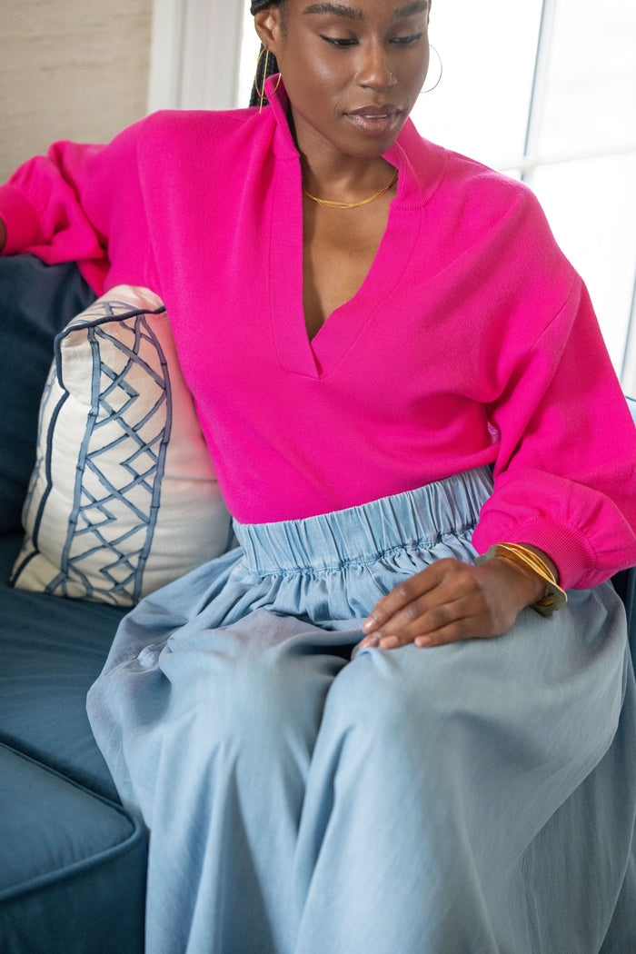 A woman sits on a blue sofa, wearing the Lolli Sweater in bright pink with blouson sleeves and a light blue high-waisted skirt. She leans on a white pillow featuring blue embroidery and accessorizes with gold jewelry.