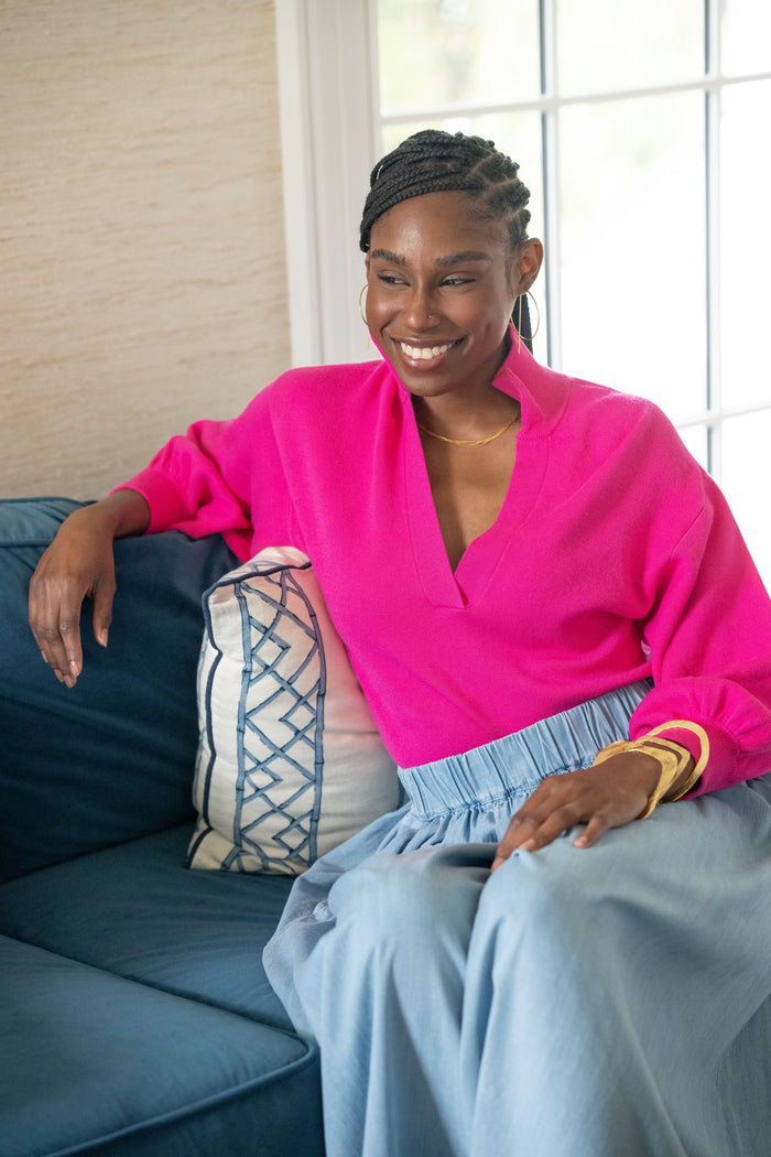 A woman with braided hair, smiling near a window, sits on a blue couch with a patterned pillow and wears the bright pink Lolli Sweater with blouson sleeves and a light blue skirt.