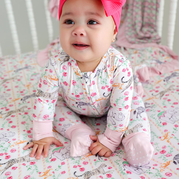 Baby in a colorful outfit sitting on a crib with a pink hat.
