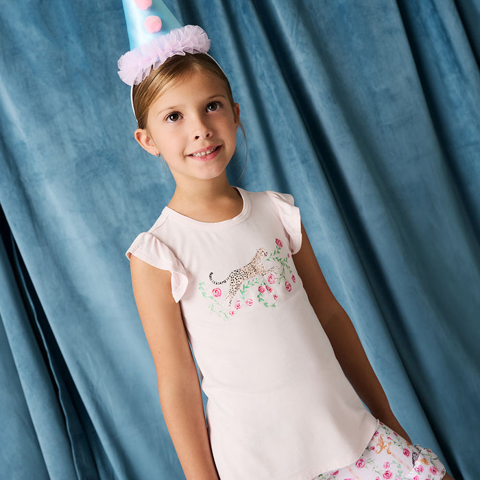 Young girl wearing a white top with floral design and a party hat against a blue curtain.