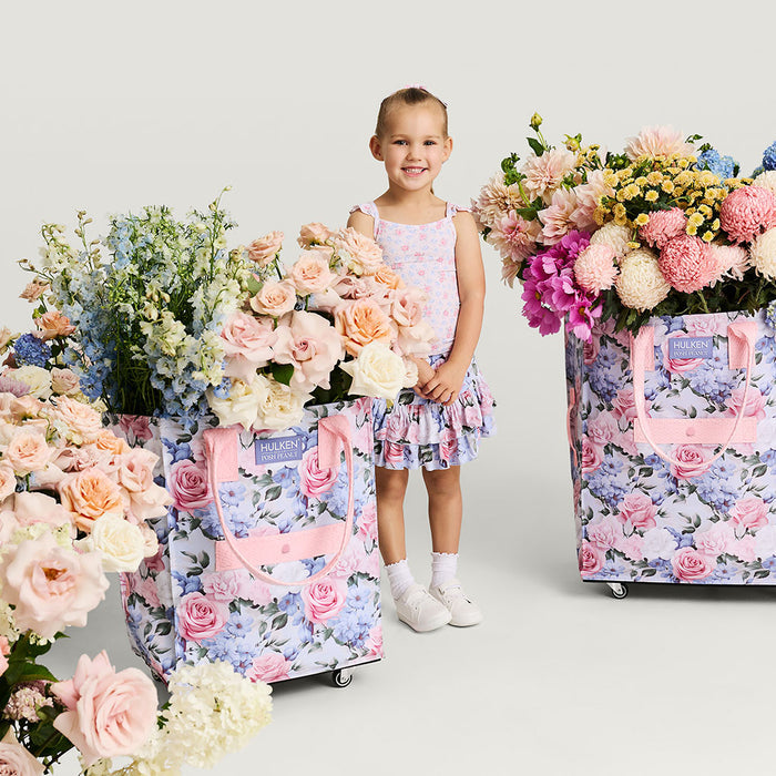 Young girl in a floral dress standing next to two large floral-patterned bags filled with flowers on a white background.