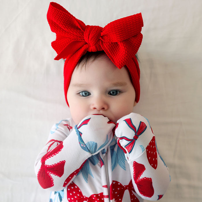 Baby wearing a red headband with a large bow and patterned mittens on a neutral background