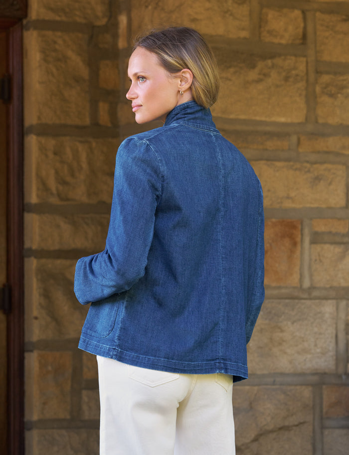 A woman wearing a blue denim jacket and cream pants, shown from the back and side profile against a tan stone wall.