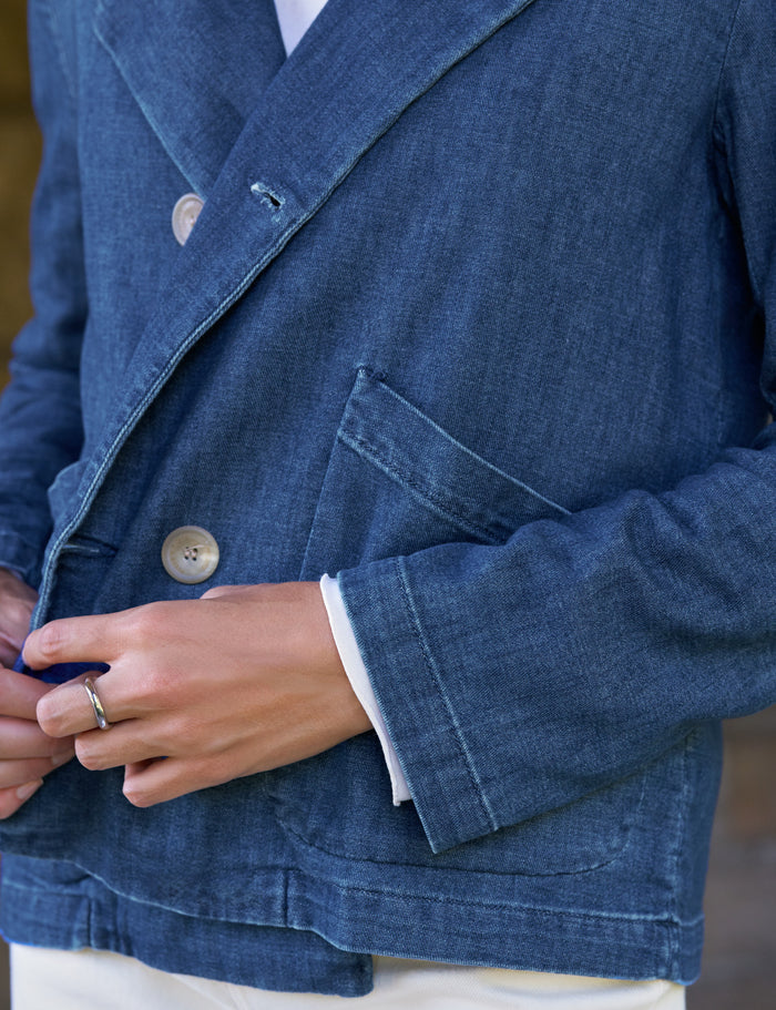 Close-up of a person wearing a blue denim-style double-breasted blazer with white shirt cuffs and a white undershirt visible, plus a silver ring on one finger.