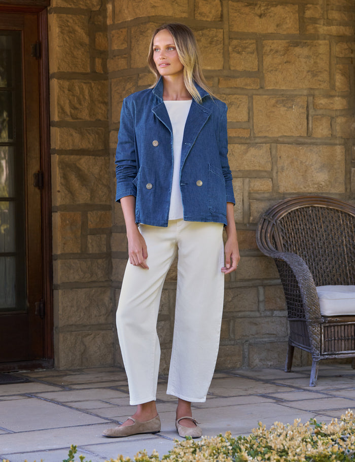A woman stands on a stone patio wearing a blue double-breasted blazer over a white top, cream cropped trousers, and tan flats. A wicker chair and tan stone wall are in the background.
