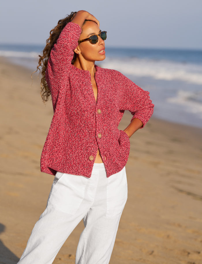 Woman in a red knitted cardigan and white pants standing on a beach.
