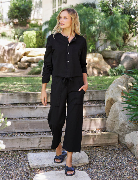 A woman wearing a black long-sleeve button-up shirt and matching black wide-leg pants stands on stone steps in a landscaped garden setting.