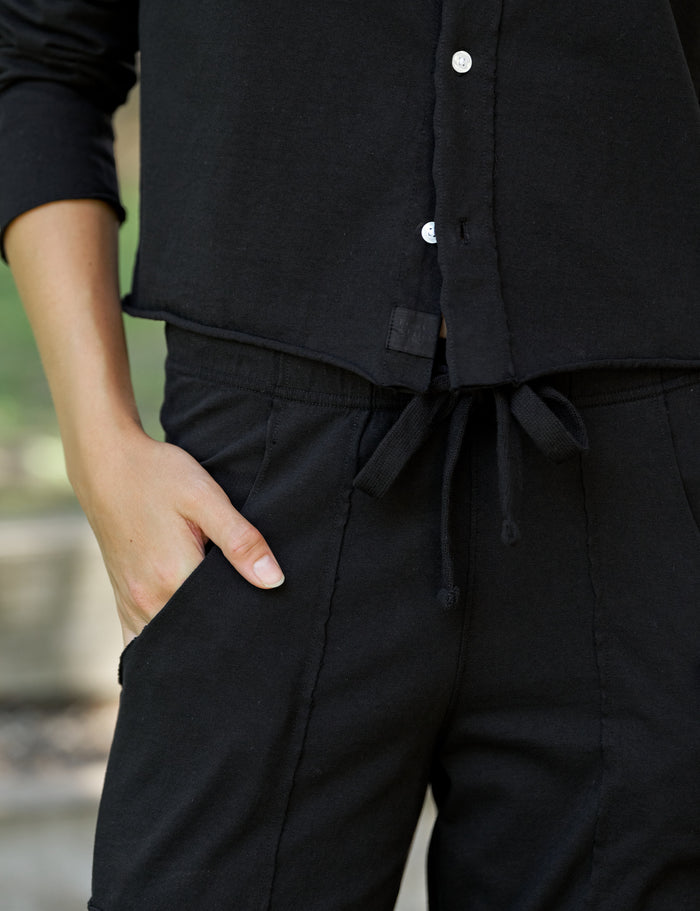 Close-up of a person wearing a black button-front top and black drawstring pants, with one hand in a pocket.