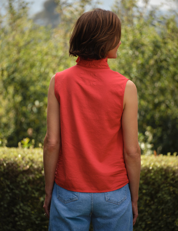 Person wearing a red sleeveless top and blue jeans standing outdoors with greenery in the background