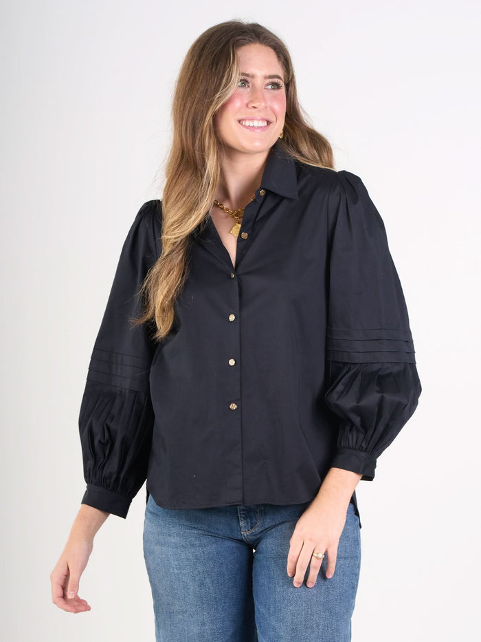 A woman with long, wavy brown hair smiles in the Oxford navy cotton poplin blouse with puffed sleeves and gold buttons, paired with blue jeans against a plain white background.