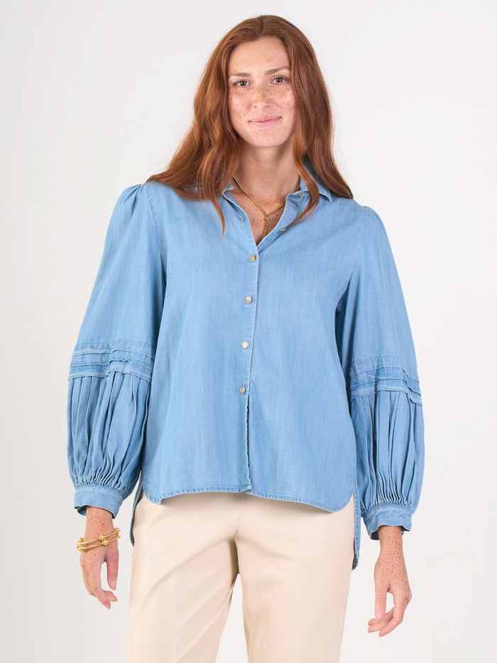 A woman with long red hair and freckles wears the Oxford denim chambray balloon sleeve blouse and cream-colored pants, standing against a plain white background.