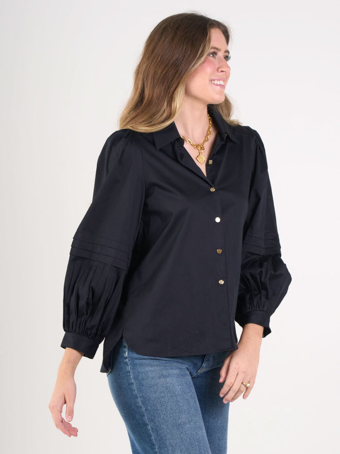 A woman with wavy light brown hair stands sideways, smiling, wearing the Oxford navy cotton poplin blouse with puffy pleated sleeves and blue jeans. She accessorizes with a gold necklace and rings.