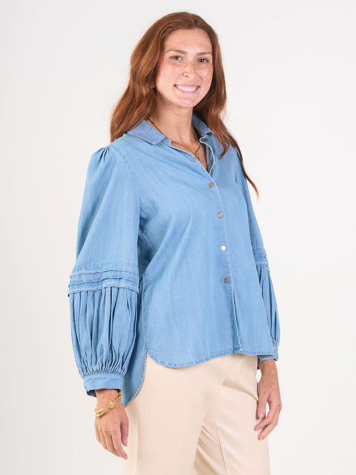 A smiling woman with long red hair wears the Oxford light blue denim chambray blouse with balloon sleeves and beige pants, standing against a plain white background.
