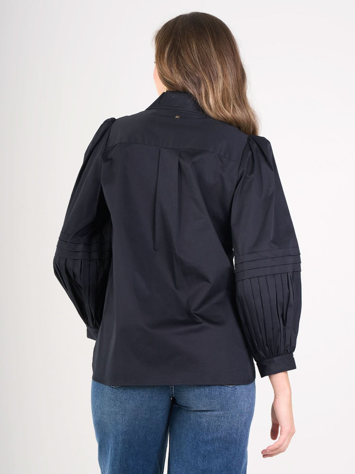 A woman with long brown hair stands with her back to the camera against a plain light background, wearing the Oxford black cotton poplin blouse with pleated balloon sleeves and blue jeans.