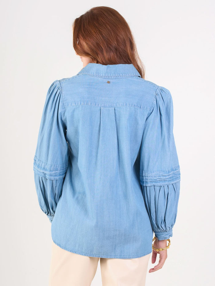 A woman with long brown hair, seen from behind, wears the Oxford light blue denim chambray shirt with balloon sleeves and beige pants, standing against a plain white background.