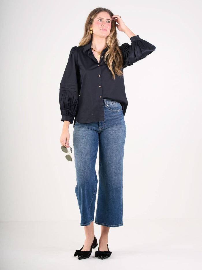Wearing the Oxford loose black cotton poplin blouse with pleated sleeves, a woman pairs it with high-waisted cropped blue jeans and black bow shoes, standing against a white background while holding sunglasses and touching her hair.