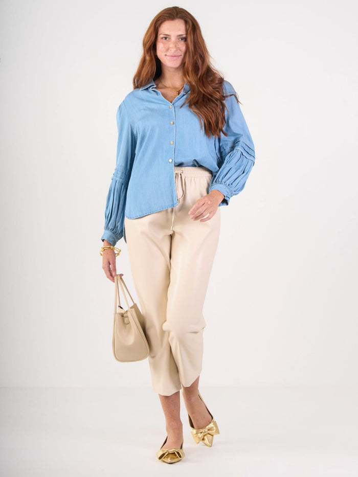 A woman with long red hair wears the Oxford light blue shirt with pleated sleeves, beige trousers, gold pointed shoes, and carries a beige handbag while smiling against a plain white background.