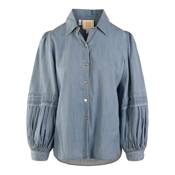 The Oxford blouse features light blue denim chambray, a collared neckline, button-down front, and long balloon sleeves with gathered detailing. Its relaxed fit and slightly curved hem give it a modern twist.