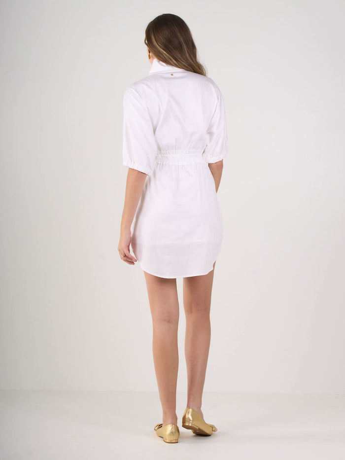 A woman with long brown hair, seen from behind, wears the Palmer Dress—a white, short-sleeved, collared style with a ruffle cinched waist—paired with gold flats against a plain white background.
