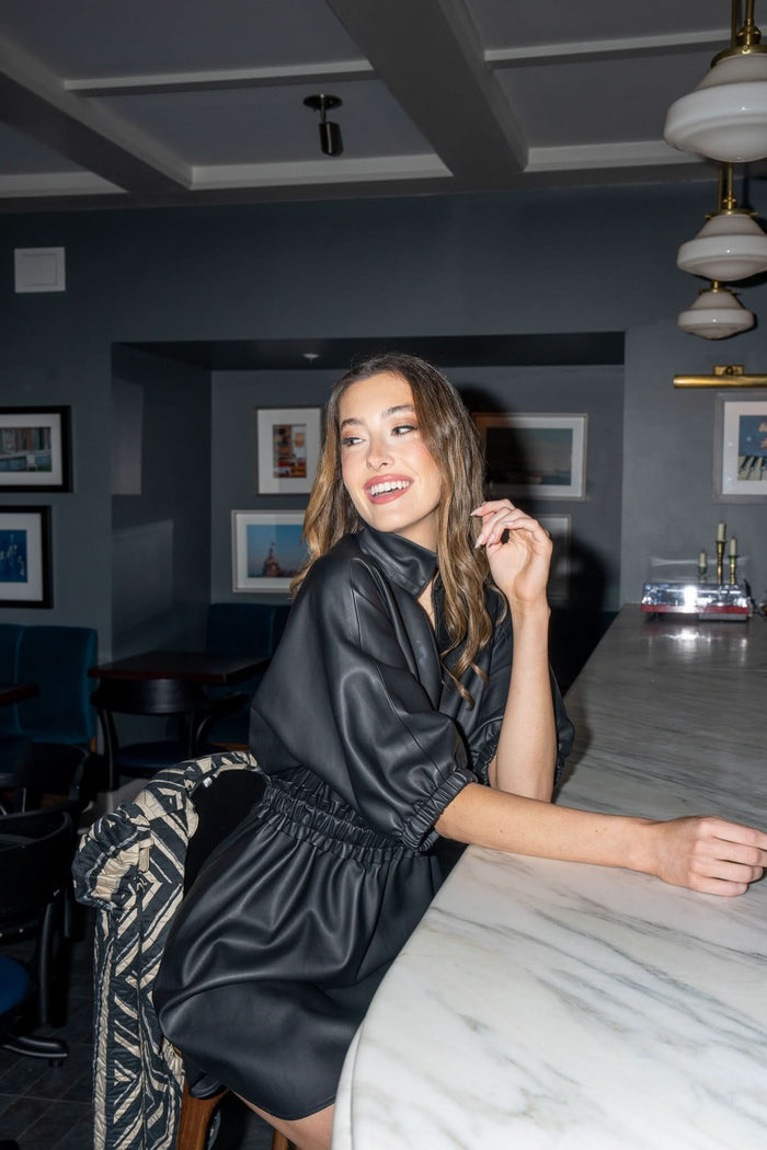 A woman in a Palmer Dress - Black Vegan Leather with a ruffle-cinched waist sits at a marble bar, smiling and looking to the side. The dark-walled room is adorned with framed pictures, and a vegan leather jacket is draped over the chair beside her.