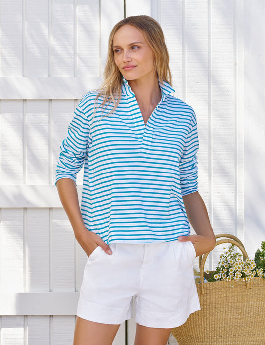 Woman wearing a blue and white striped shirt and white shorts standing against a white wooden fence.