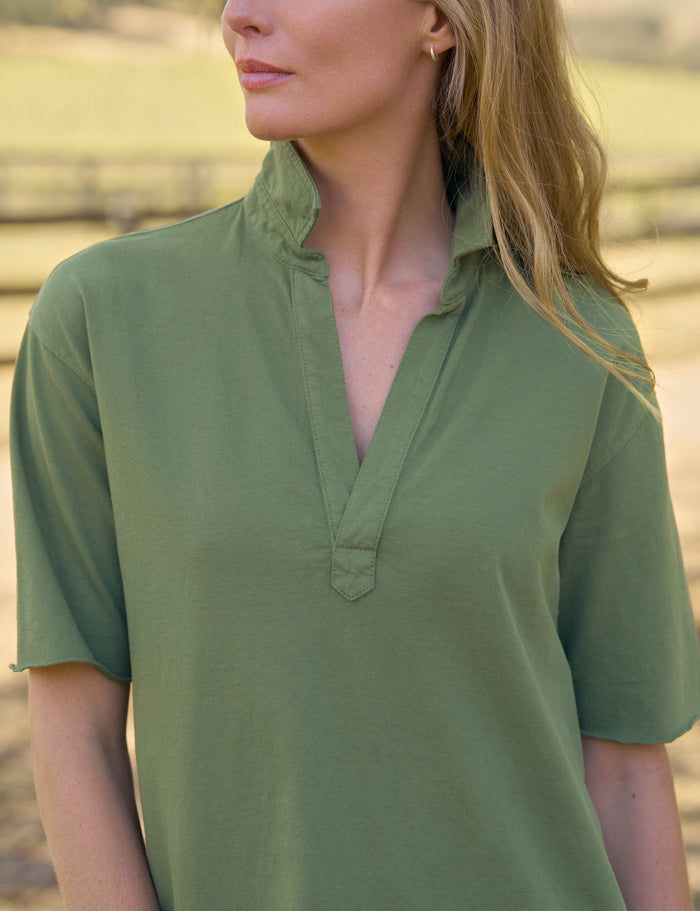 A woman wearing a short-sleeve olive green shirt with a split collar, photographed outdoors with a blurred natural background.