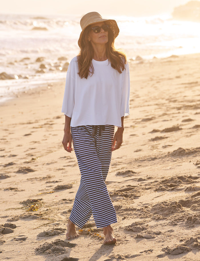 Woman in white top and striped pants standing on a sandy beach with sunset in the background