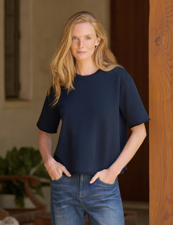 A woman with blonde hair wearing a dark navy blue short-sleeve top and blue jeans, standing in front of a wooden door.