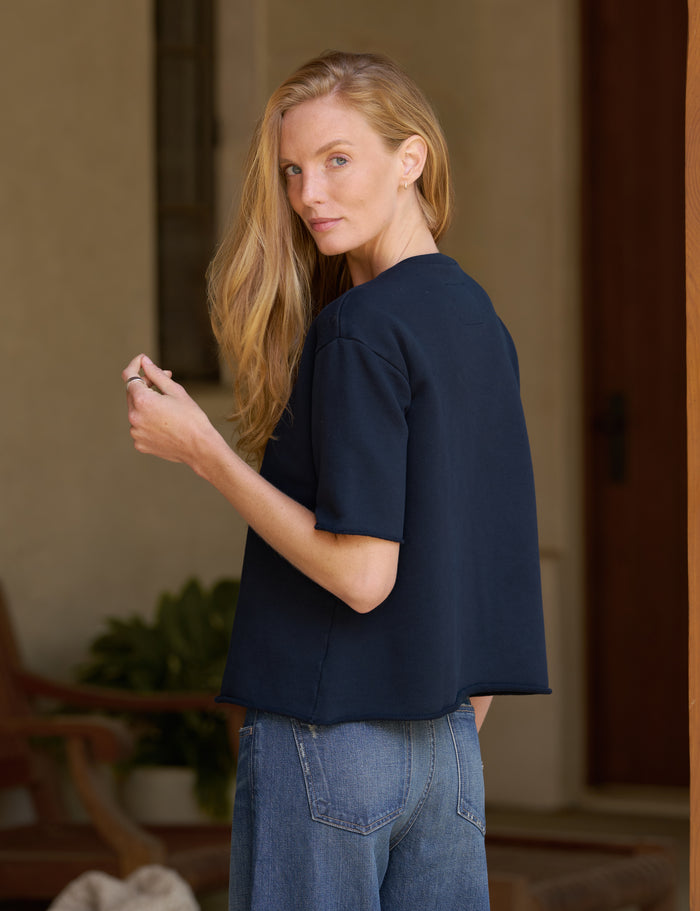 A woman with blonde hair is shown from the back wearing a dark navy blue short-sleeve top and blue jeans, standing in a warm-toned indoor setting.