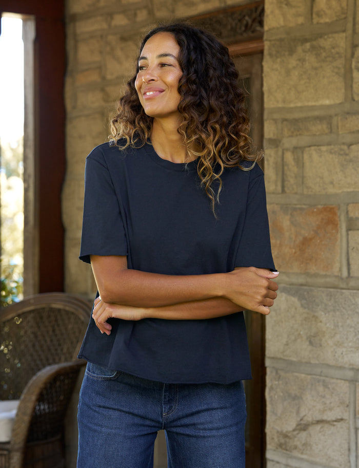 A woman with curly brown hair wearing a dark navy blue short-sleeve t-shirt and blue jeans, standing on a stone porch with her arms crossed.