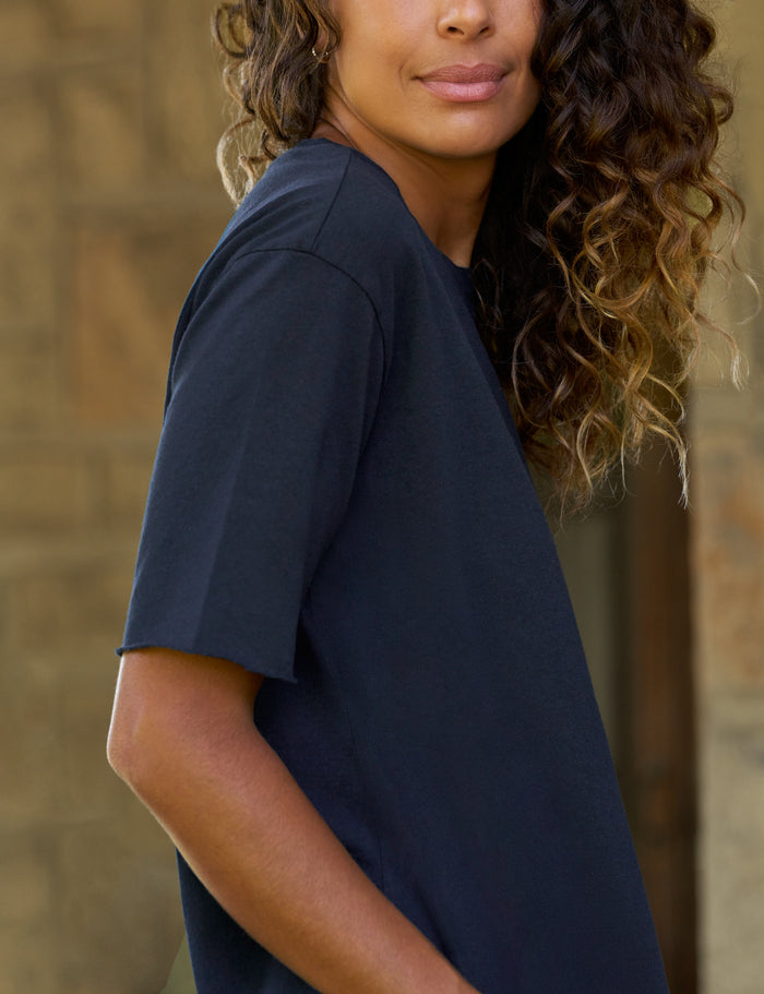A woman wearing a dark navy blue short-sleeve t-shirt, shown from a side-back angle against a blurred stone wall background.