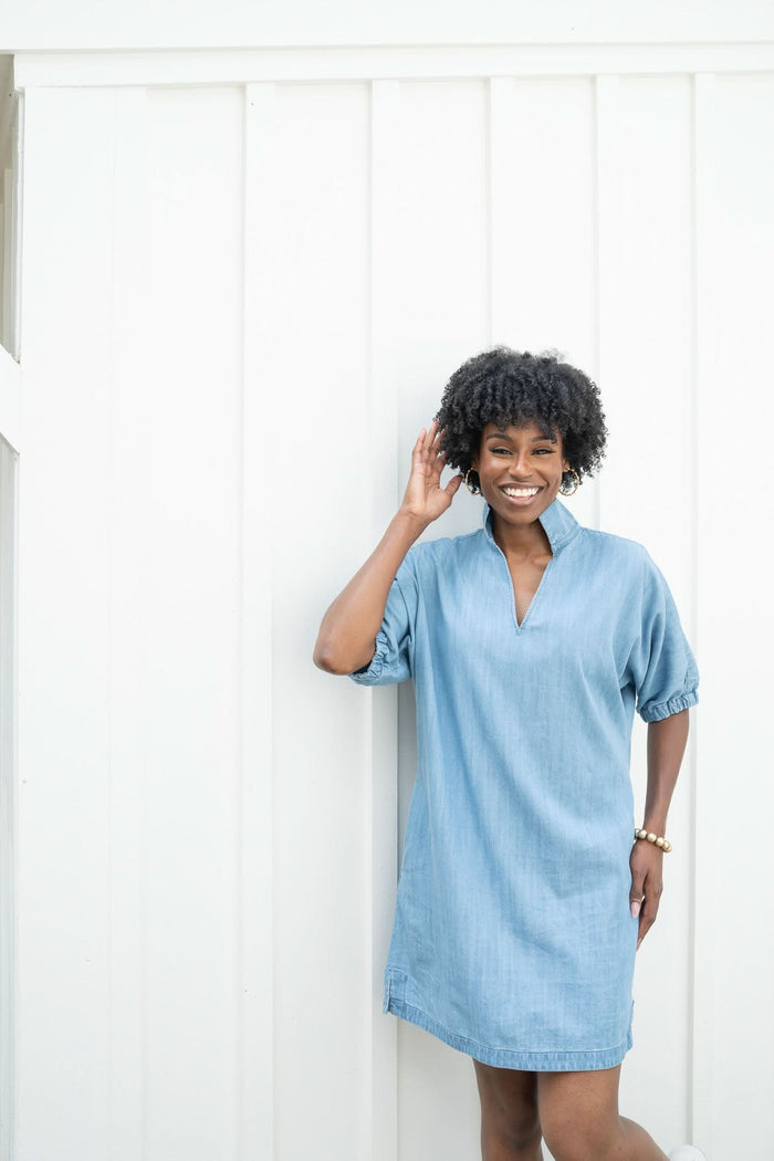 Wearing the Poppy Dress in light blue denim chambray, a person stands smiling with one hand touching their hair against a white wall. Accessorized with a bracelet, this dress showcases a v-neck and elbow-length sleeves, creating an atmosphere that is both bright and cheerful.
