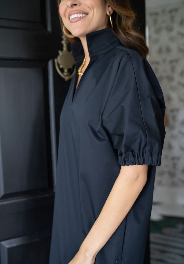 A woman wearing the Poppy Dress in black cotton poplin by Emily McCarthy Clothing, featuring a high collar and short puff sleeves, stands near a dark wooden door. She is accessorized with gold hoop earrings and a necklace, smiling gently against the backdrop of floral-patterned wallpaper.