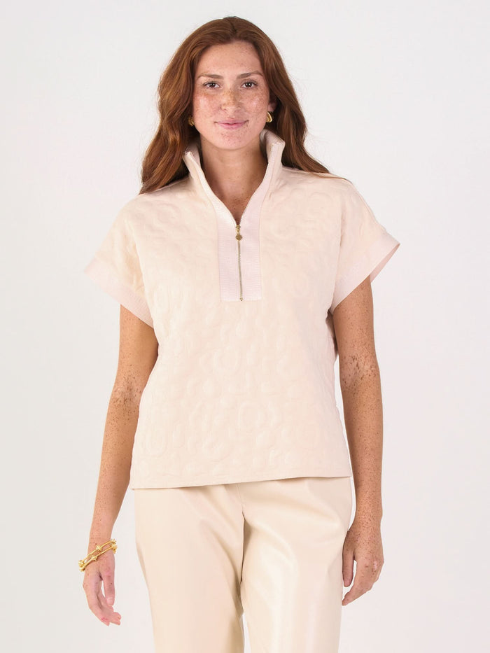A woman with long red hair wears the Poppy Pullover in light beige, featuring a 3/4 zip collar, paired with matching beige pants. She accessorizes with a gold bracelet and necklace against a plain light background.