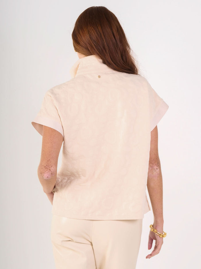 A woman with long red hair, seen from behind, wears a short-sleeved, light beige Poppy Pullover in embossed cheetah fabric with a high collar and matching pants. She accessorizes with a gold bracelet on her right wrist.