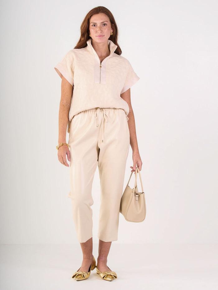 A woman with long red hair models the Poppy Pullover, a light beige, short-sleeved zip-up top paired with matching embossed cheetah pants. She accessorizes with a beige handbag, gold flats, and gold jewelry against a white background.