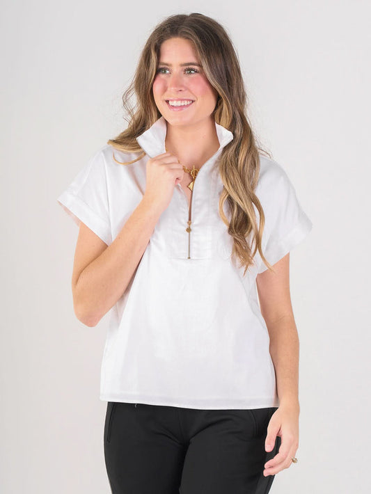 A smiling woman with long wavy hair wears the white Poppy Pullover, a short-sleeve cotton poplin top with a half-zipper, paired with black pants against a plain light background—highlighting elevated basics.