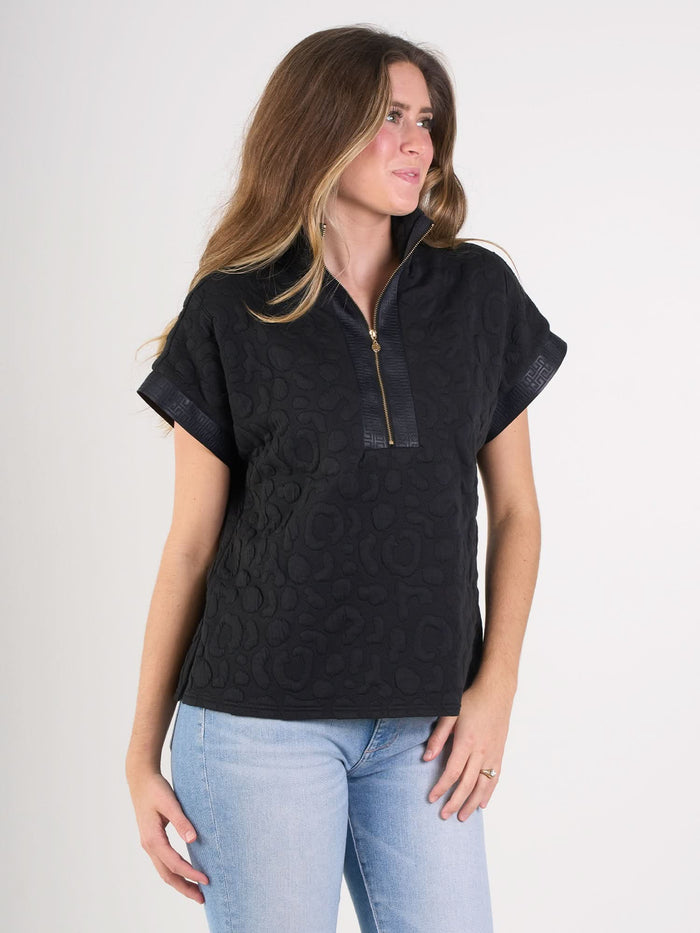 A woman with long brown hair wears the Poppy Pullover, a textured black short-sleeve top with a zippered collar, paired with light blue jeans against a white background, creating a sporty and sophisticated look.
