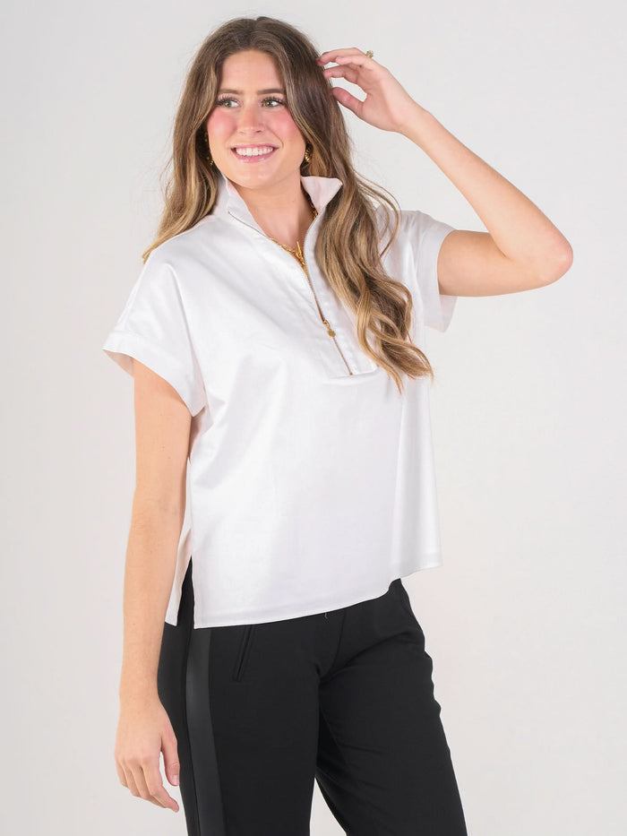 A woman with long, wavy hair smiles and poses with one hand raised, wearing the short-sleeved white cotton poplin Poppy Pullover and black pants against a plain light background, showcasing elevated basics.