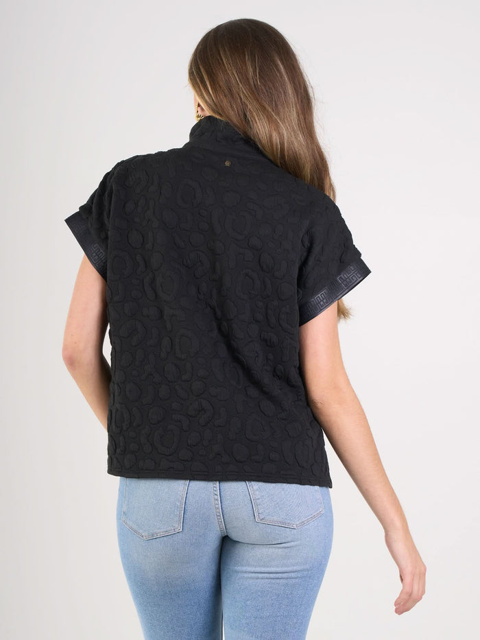 A woman with long brown hair, wearing the Poppy Pullover in Black Cheetah texture and light blue jeans, stands with her back to the camera against a plain white background, showcasing sporty sophistication.