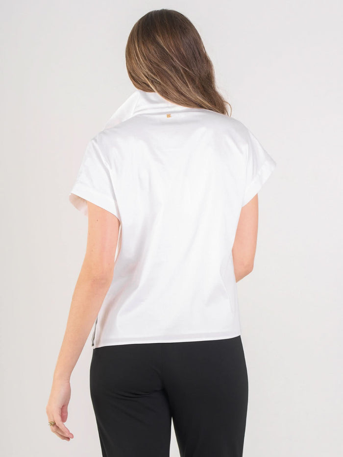 A woman with long brown hair faces away, wearing the short-sleeved white Poppy Pullover and black pants against a plain light background.