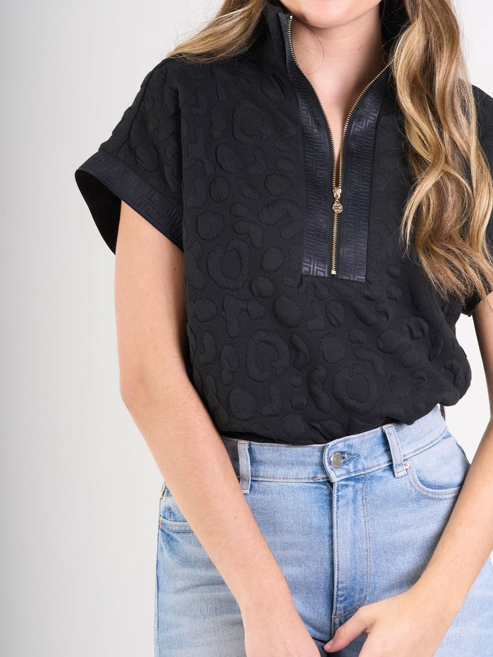 A woman wears the Poppy Pullover, a textured black cheetah short-sleeve top with a zip-up collar, paired with light blue high-waisted jeans for a sporty yet sophisticated look against a plain light background.