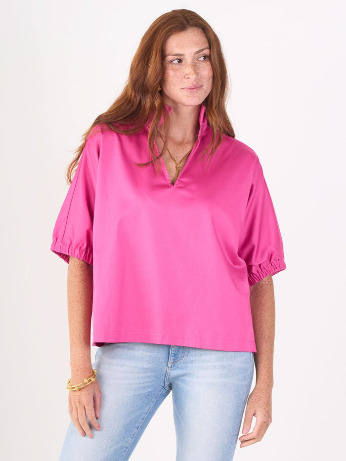 A woman with long red hair wears the Poppy Top and light blue jeans with a stylish fuchsia fedora, standing against a plain white background.