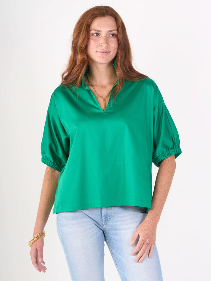 A woman with long red hair and freckles wears the bright green Poppy Top in airy cotton poplin with a V-neck, paired with light blue jeans and gold jewelry, standing against a plain white background.